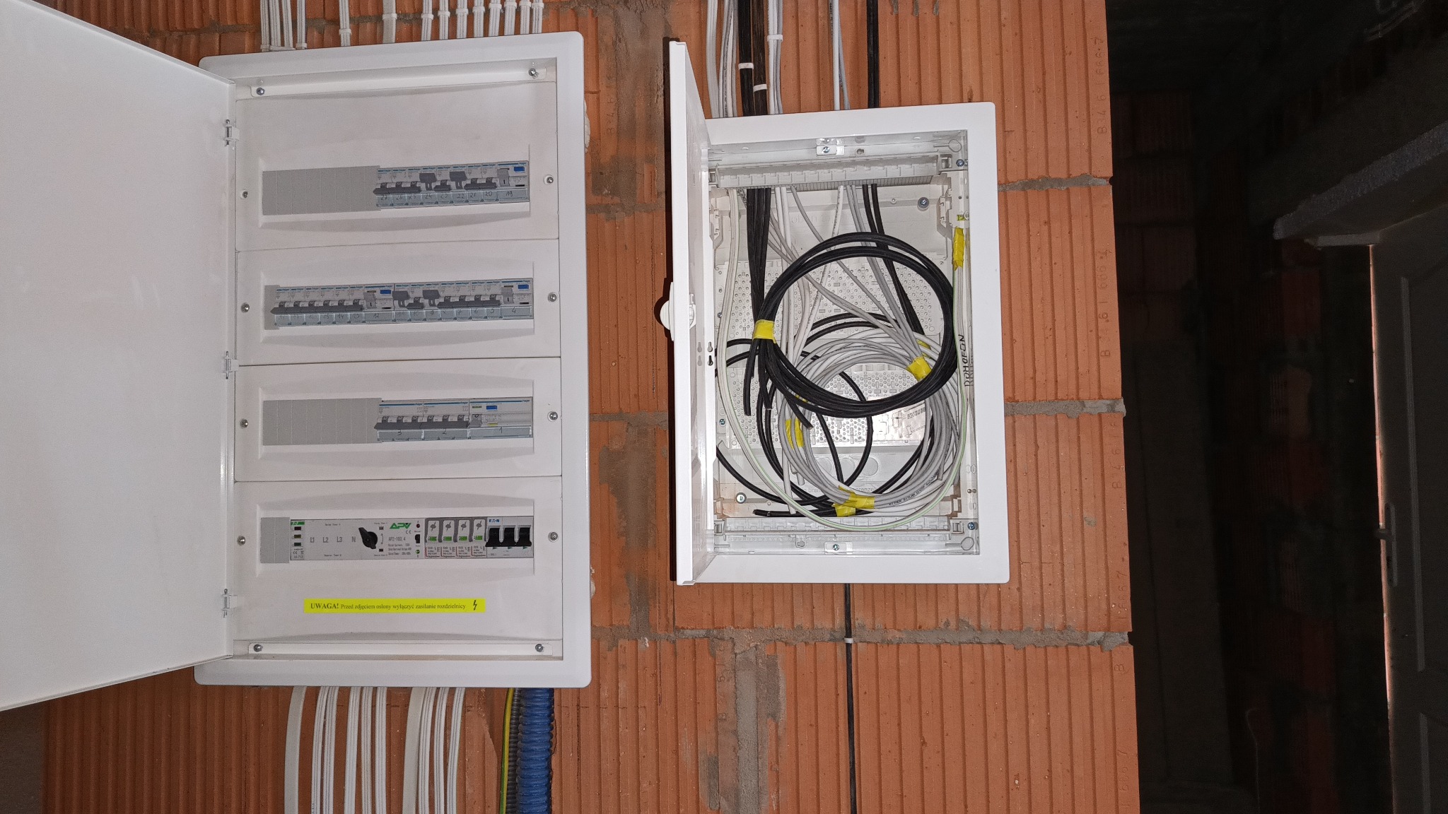 Dwie białe skrzynki elektryczne na ceglanej ścianie. Jedna otwarta, z widocznymi kablami, druga z bezpiecznikami i żółtą naklejką ostrzegawczą. Instalacja w trakcie realizacji.