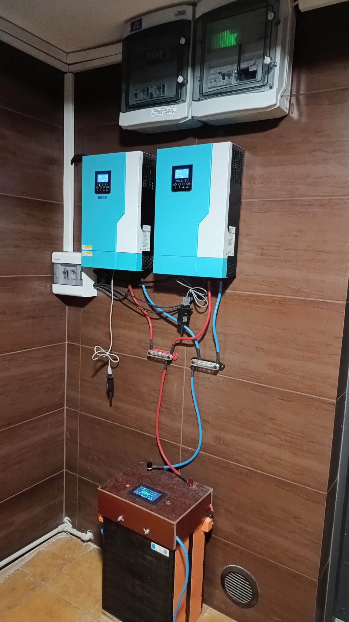 Kompletna instalacja fotowoltaiczna w pomieszczeniu z brązowymi płytkami, widoczne inwertery, rozdzielnice i akumulator z podłączeniami kablowymi.