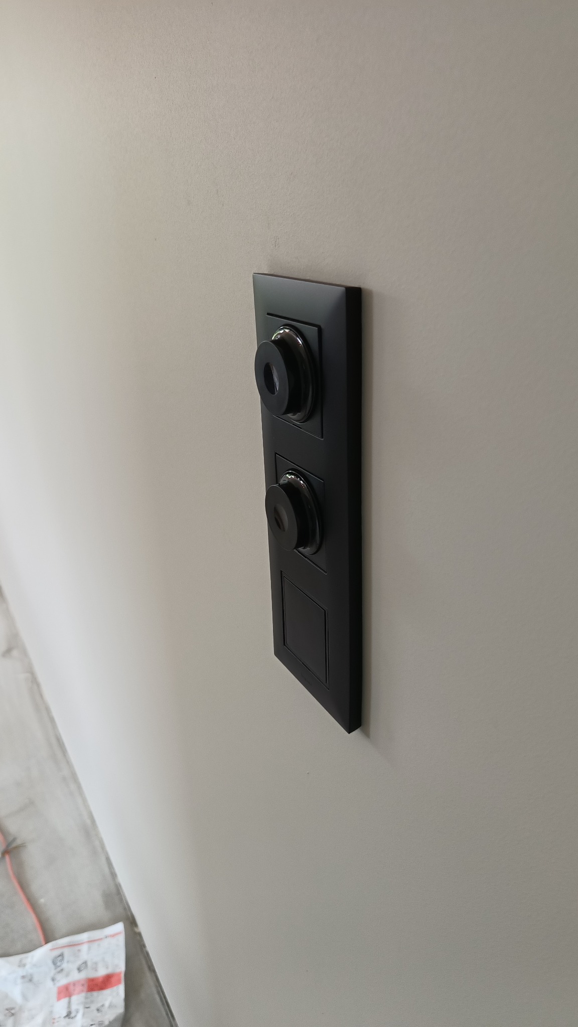 Czarny panel sterowania oświetleniem z dwoma okrągłymi przyciskami i prostokątnym elementem na beżowej ścianie. Minimalistyczny design, nowoczesne wykończenie.