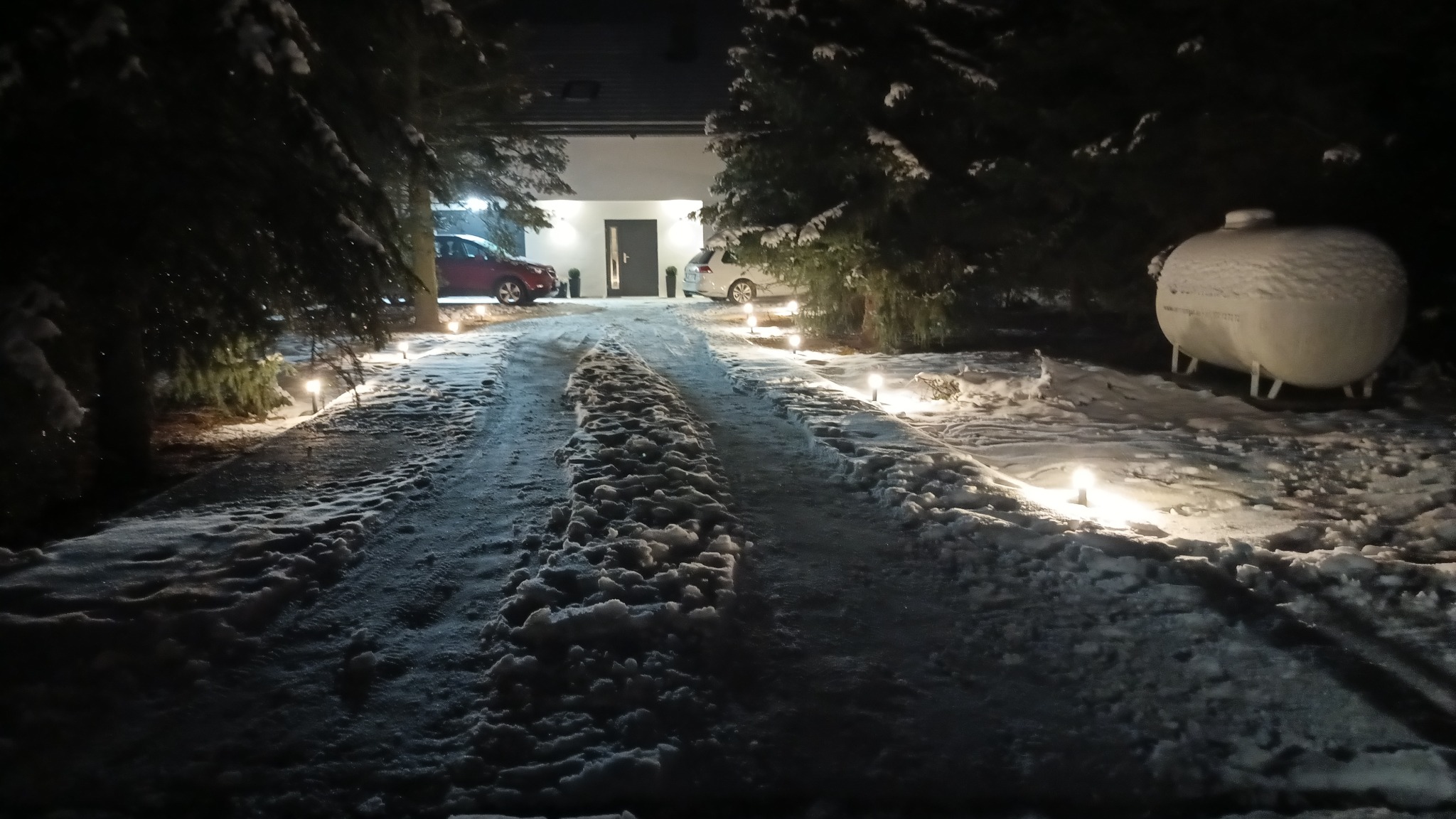 Zimowy krajobraz posesji z podjazdem oświetlonym lampami najazdowymi. W tle dom z oświetlonym wejściem i zaparkowanymi samochodami. Po prawej zbiornik na gaz.
