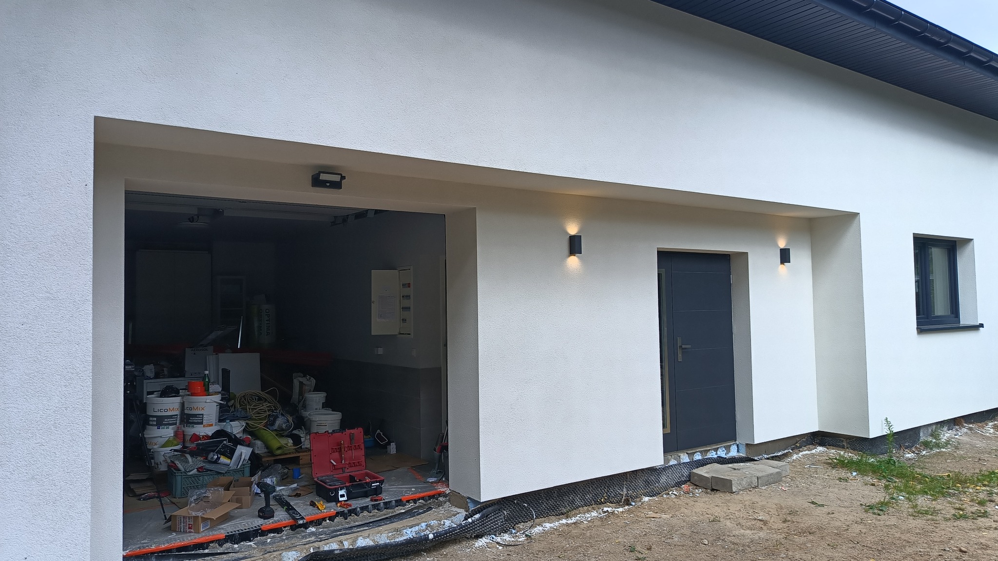 Elewacja budynku z zamontowanym oświetleniem zewnętrznym. Otwarta brama garażowa z widocznymi narzędziami i materiałami budowlanymi wewnątrz.