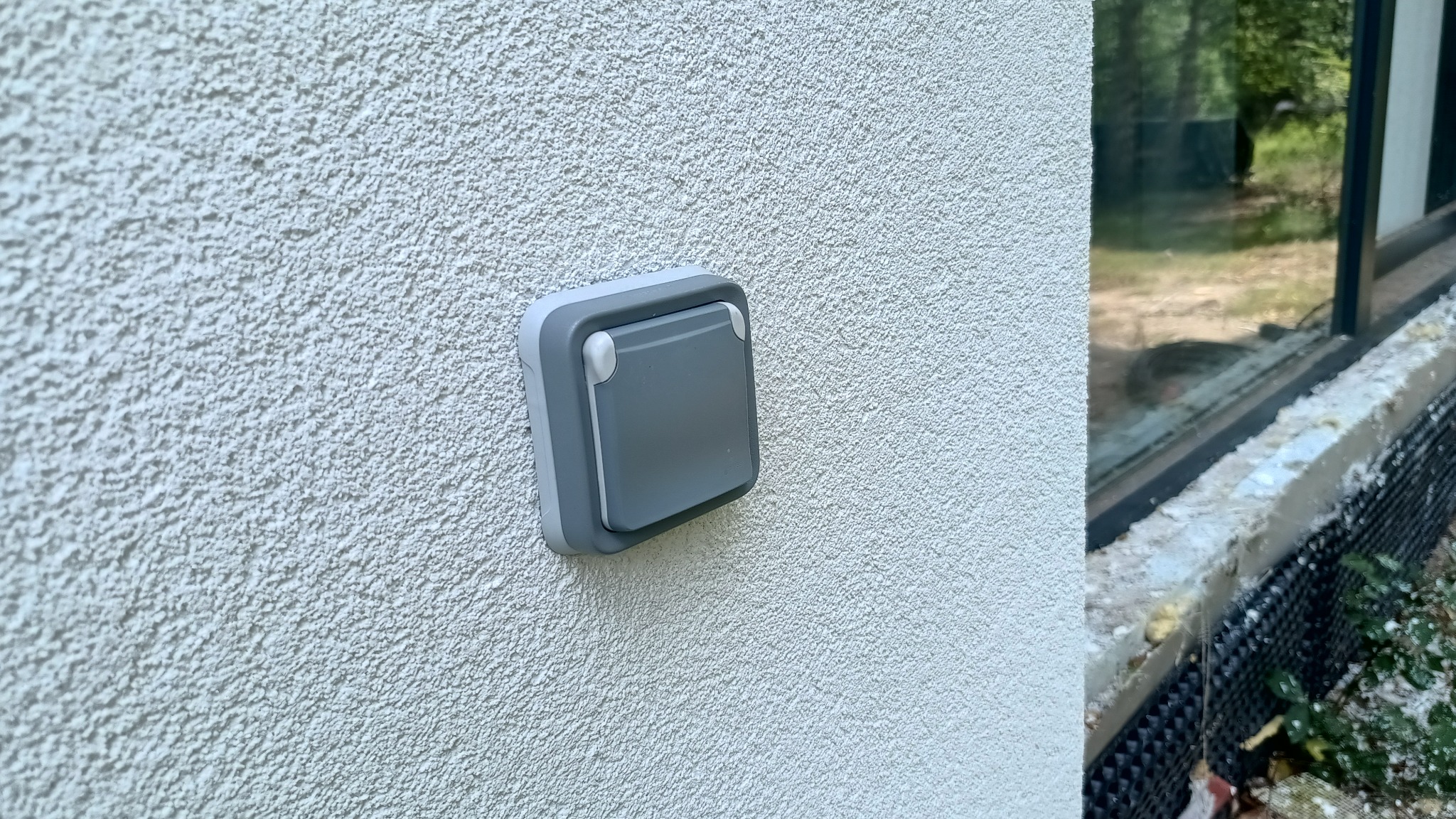 Szary, hermetyczny wyłącznik na elewacji budynku o grubej fakturze, z widocznym oknem w tle. Detal instalacji elektrycznej zewnętrznej.