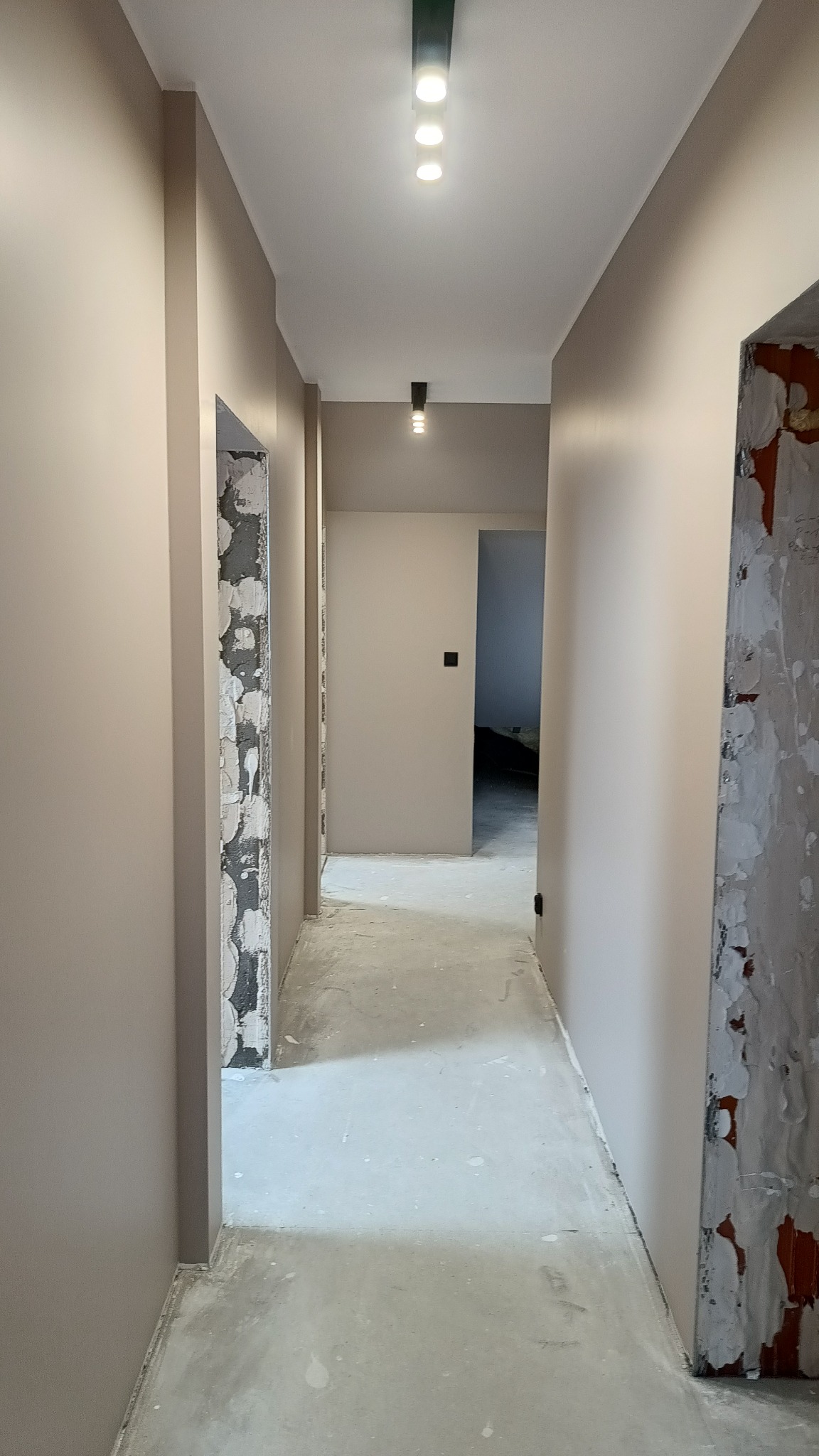 Korytarz w trakcie remontu: świeżo pomalowane ściany w odcieniach beżu, widoczne fragmenty surowych ścian przy ościeżnicach, nowoczesne oświetlenie sufitowe.