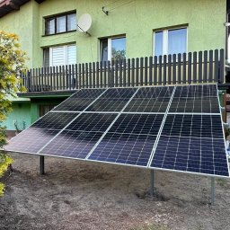AYON ENERGY SPÓŁKA Z OGRANICZONĄ ODPOWIEDZIALNOŚCIĄ - Instalacja fotowoltaiczna na trawniku przed zielonym domem z balkonem. Panele ustawione pod kątem, widoczne podpory. W tle altana i osoby przy stole.