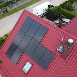 AYON ENERGY SPÓŁKA Z OGRANICZONĄ ODPOWIEDZIALNOŚCIĄ - Czerwony dach z fotowoltaiką w kształcie litery L, komin z anteną satelitarną, okno dachowe. Widok z góry na dom, ogród i ulicę z pojazdami.