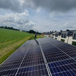 AYON ENERGY SPÓŁKA Z OGRANICZONĄ ODPOWIEDZIALNOŚCIĄ - Instalacja fotowoltaiczna na dachu domu jednorodzinnego z widokiem na zielone pole i zabudowania. Monter stoi na panelach.