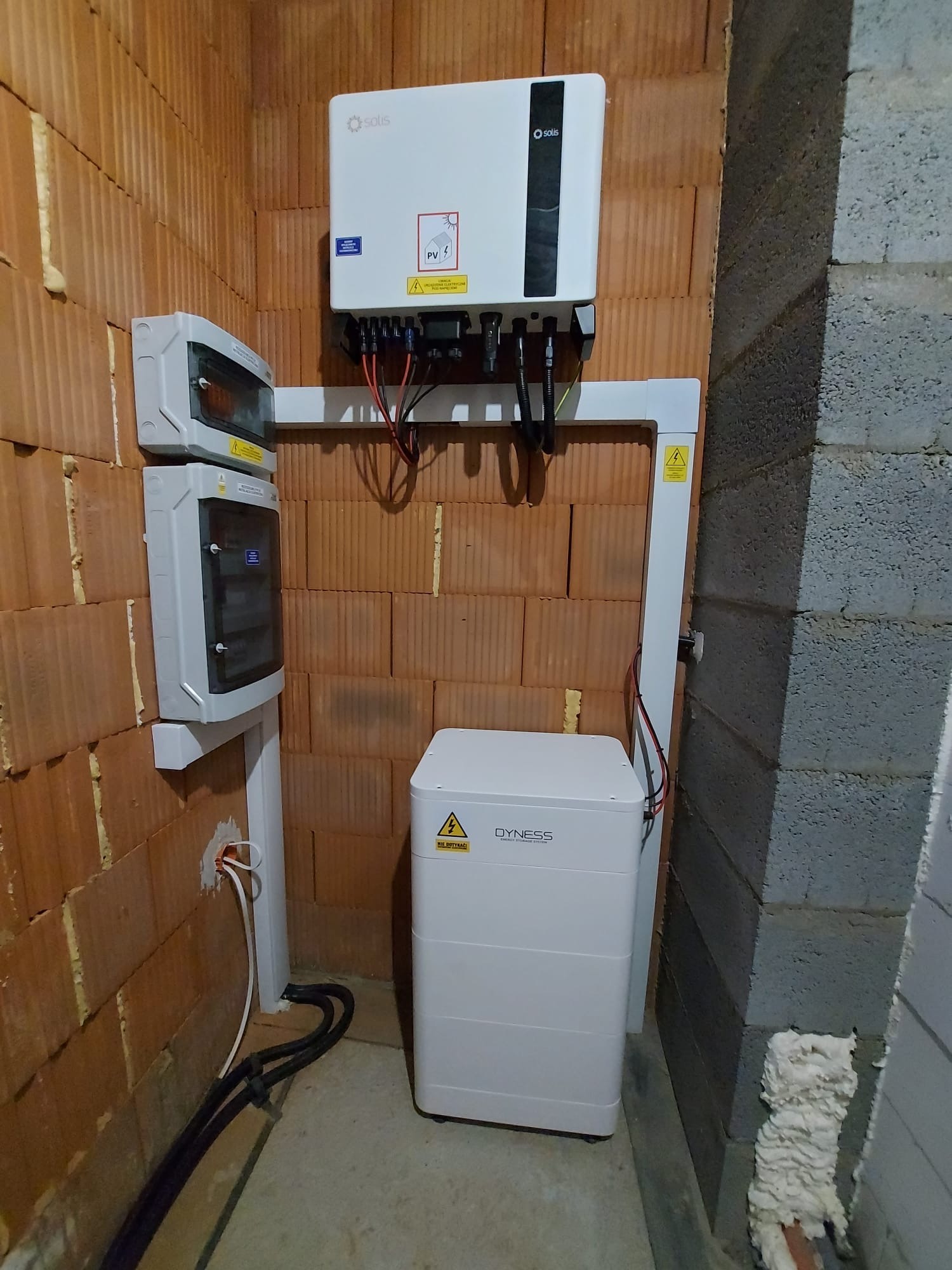 Instalacja fotowoltaiczna w pomieszczeniu: inwerter Solis, biały magazyn energii Dyness i rozdzielnica na ścianie z czerwonej cegły. Widoczne przewody i rury ochronne.