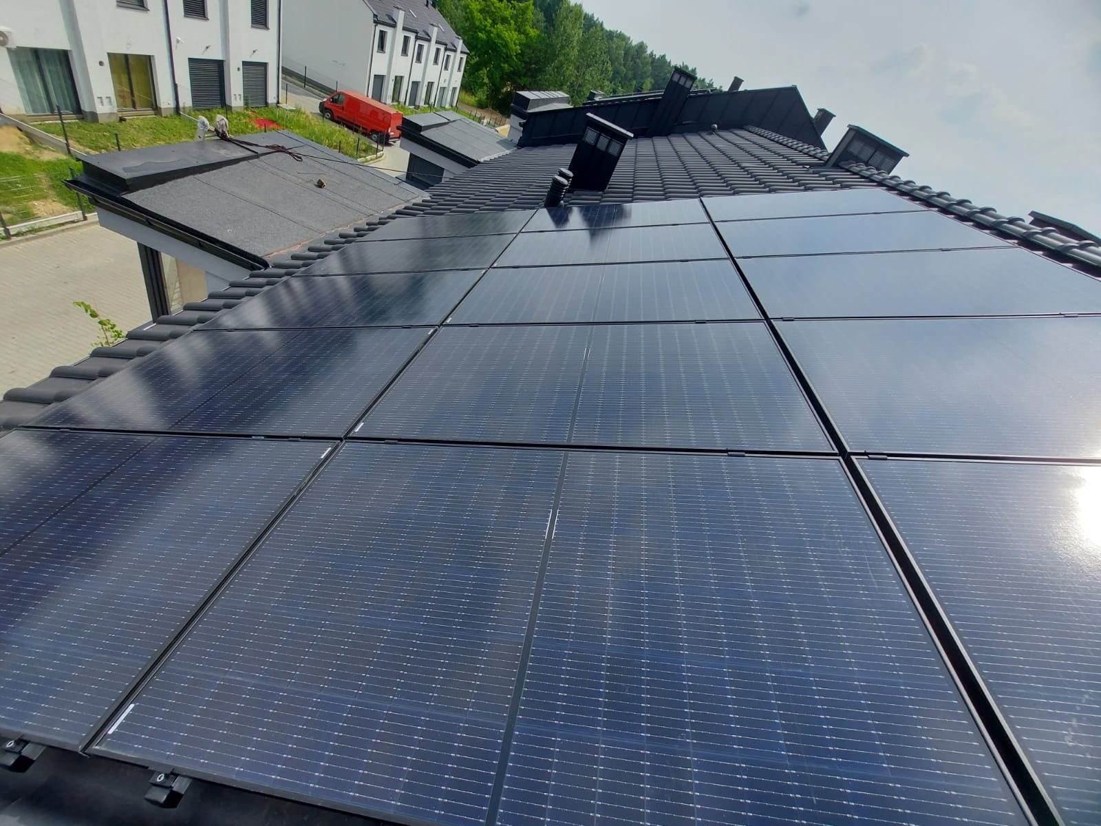 Panele fotowoltaiczne na dachu domu jednorodzinnego z ciemną dachówką. Widok z góry, w tle budynki i zieleń.