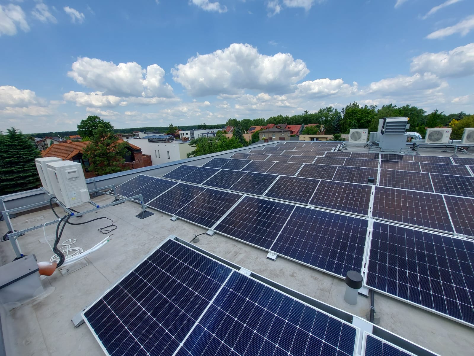 Instalacja fotowoltaiczna na dachu budynku z klimatyzacją, widok z góry, błękitne niebo z chmurami w tle. Panele w regularnych rzędach, widoczne elementy montażowe.