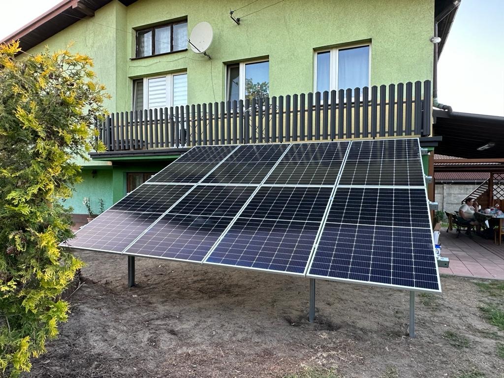 Instalacja fotowoltaiczna na trawniku przed zielonym domem z balkonem. Panele ustawione pod kątem, widoczne podpory. W tle altana i osoby przy stole.