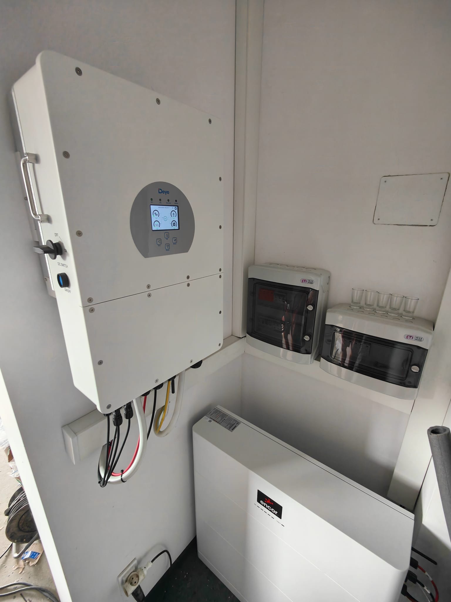 Wnętrze pomieszczenia z zamontowanym inwerterem Deye, akumulatorem Encor i rozdzielnicami ETI, okablowanie widoczne, instalacja w białym pomieszczeniu.