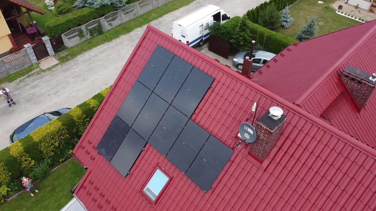 Czerwony dach z fotowoltaiką w kształcie litery L, komin z anteną satelitarną, okno dachowe. Widok z góry na dom, ogród i ulicę z pojazdami.