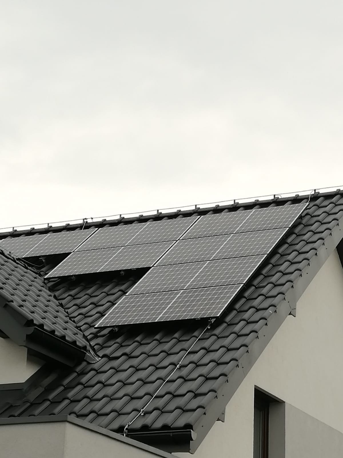 Panele fotowoltaiczne na dachu domu z czarną dachówką, widoczne przewody i elementy montażowe. Pochmurne niebo w tle. Ujęcie z dołu, lekko z boku.
