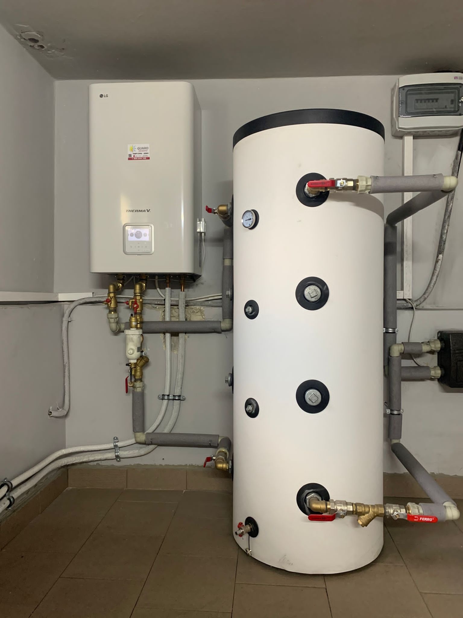 Biały zbiornik akumulacyjny ciepłej wody użytkowej z zaworami Ferro i wiszący moduł LG Therma V, połączone rurami w szarej kotłowni z brązowymi płytkami podłogowymi.