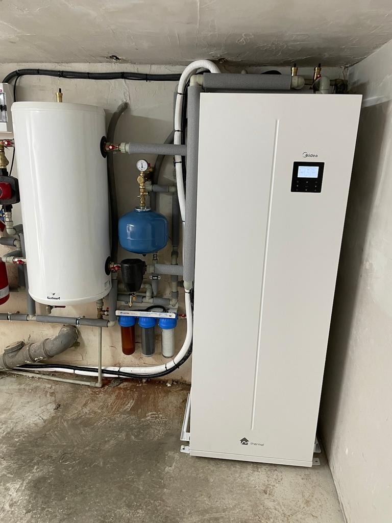 Biała pompa ciepła Midea z zbiornikiem Bułmet i filtrami wody. Instalacja grzewcza w piwnicy z szarymi rurami i betonową podłogą.