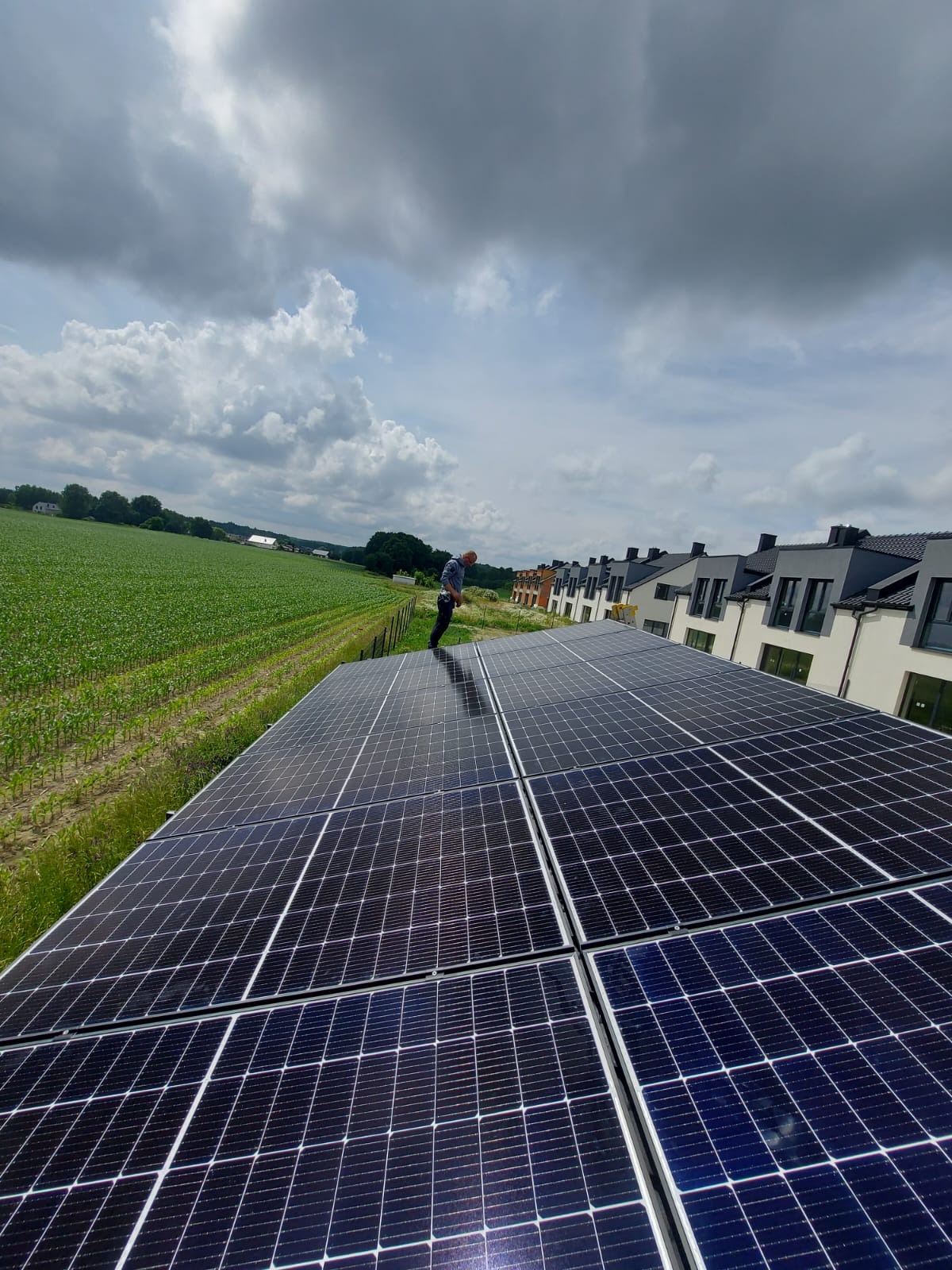 Instalacja fotowoltaiczna na dachu domu jednorodzinnego z widokiem na zielone pole i zabudowania. Monter stoi na panelach.