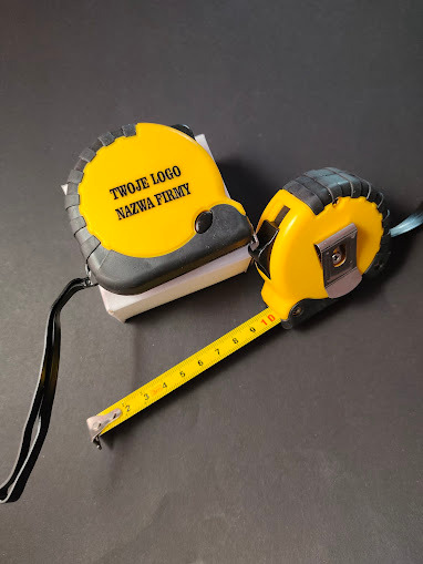 Dwie żółte miarki zwijane z czarnymi elementami. Na większej nadruk: Twoje Logo Nazwa Firmy. Gadżety reklamowe z logo firmy na ciemnym tle.