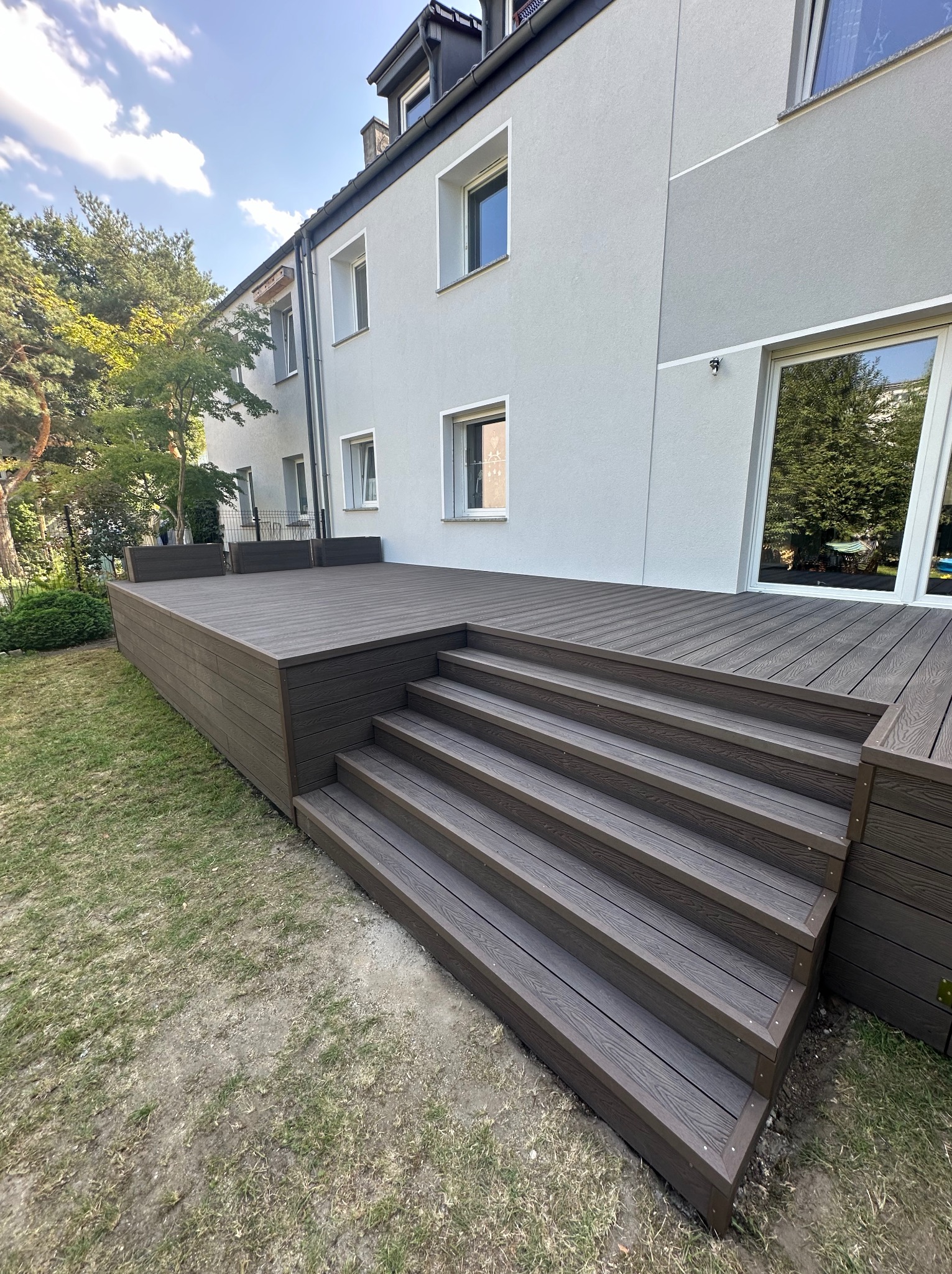 Nowoczesny taras kompozytowy z brązowymi deskami i schodami w Warszawie, przylegający do jasnoszarego budynku z dużymi oknami.