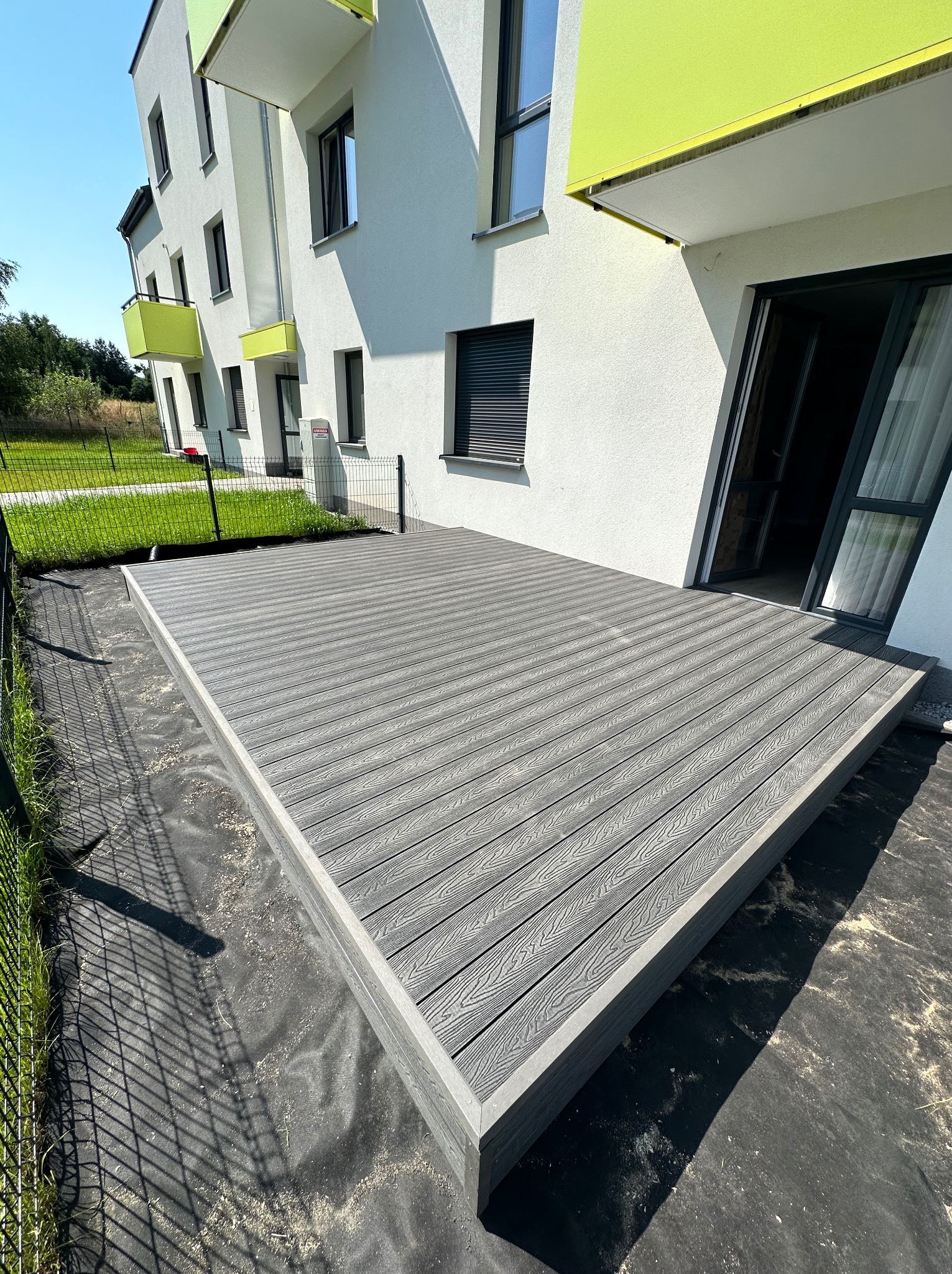 Szara deska tarasowa z kompozytu na przygotowanym podłożu, widok z boku budynku z zielonymi balkonami i czarną siatką ogrodzeniową.