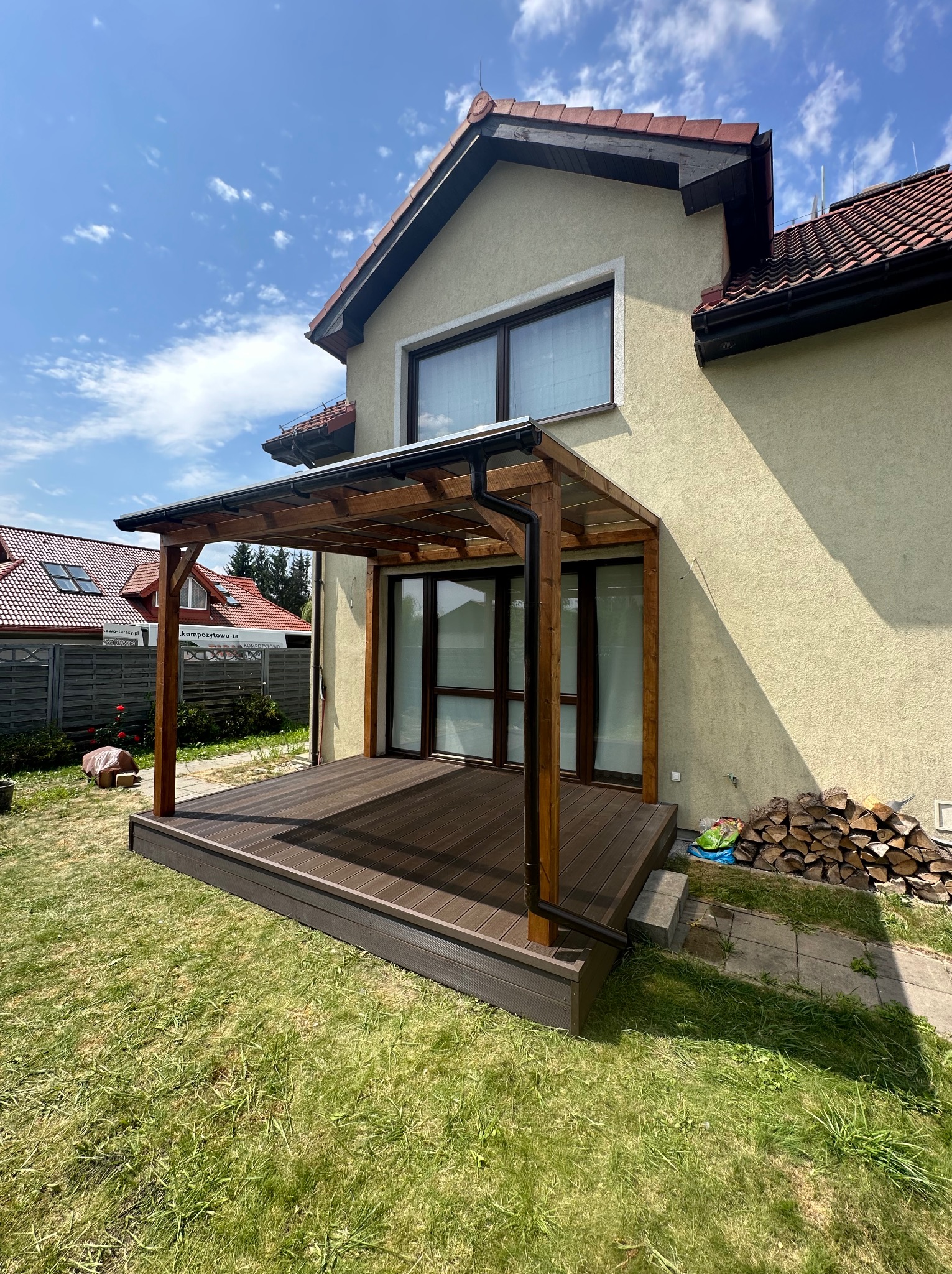 Nowoczesny taras kompozytowy z drewnianą pergolą przylegający do domu, widok z trawnika w słoneczny dzień, z widocznym stosem drewna opałowego.