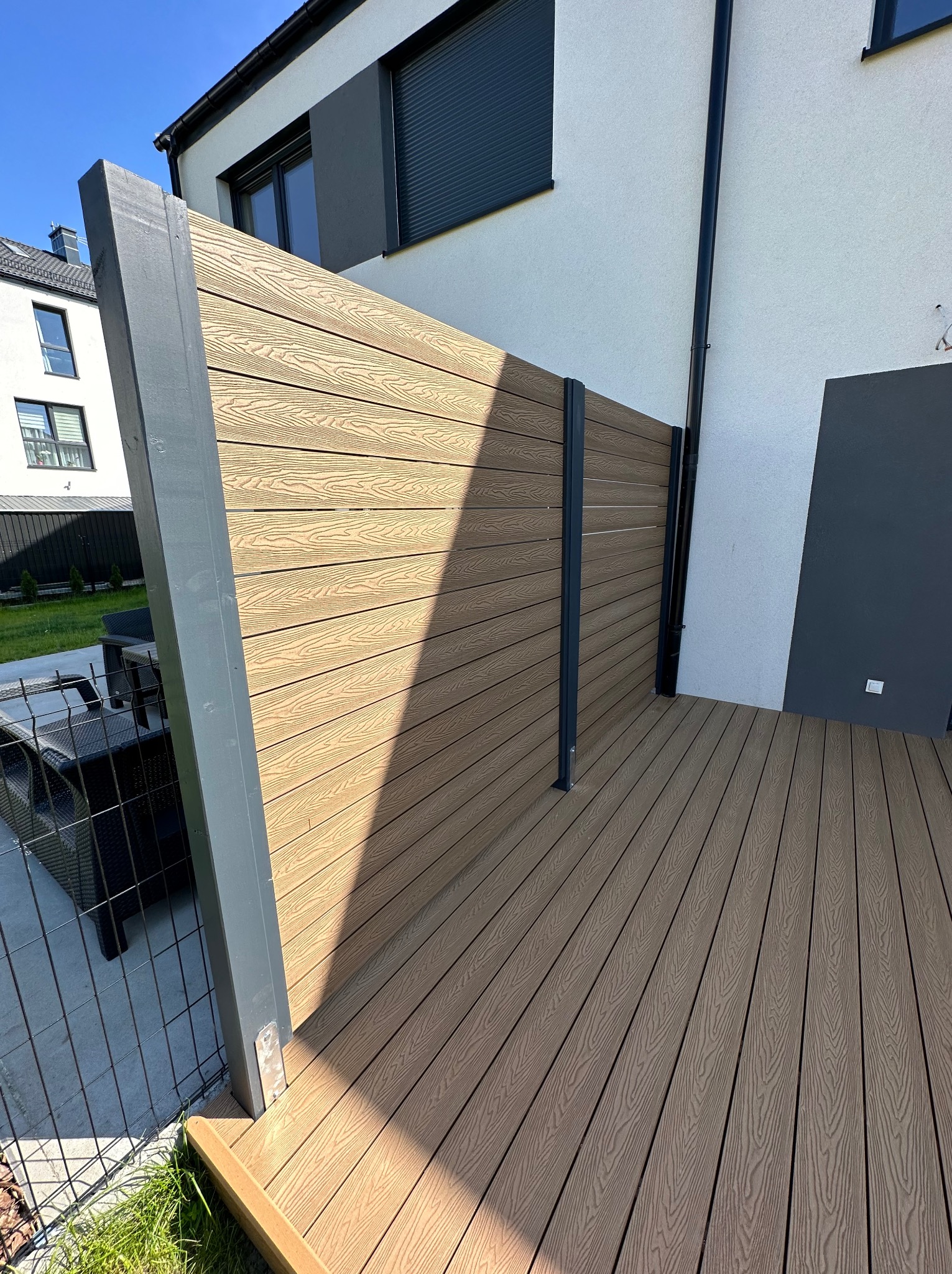 Nowoczesny taras z desek kompozytowych i panelu ogrodzeniowego w kolorze drewna, z szarymi słupkami, przylegający do białego budynku z szarą rynną.