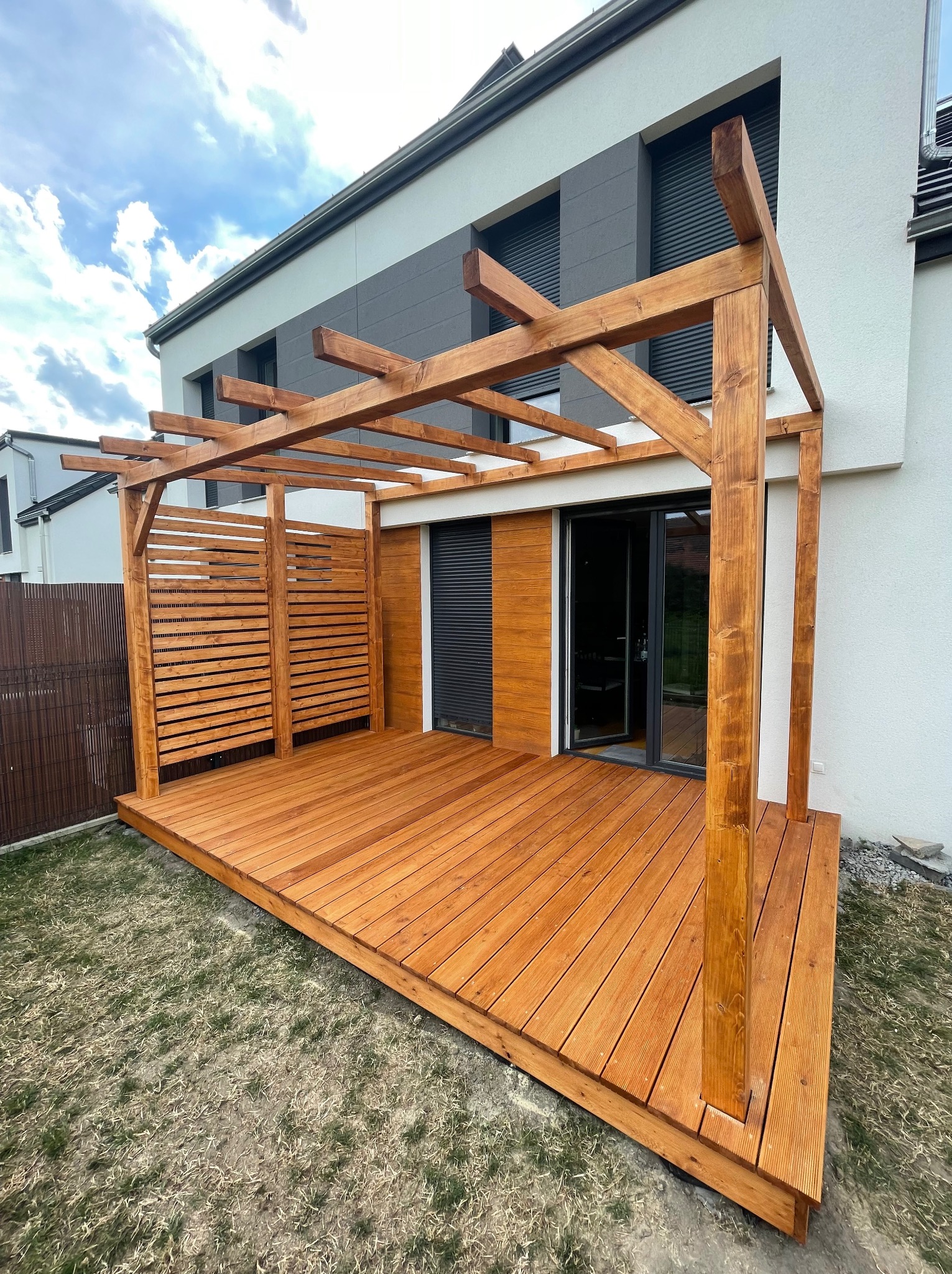 Nowoczesny taras z jasnego drewna z pergolą przylegający do domu z szarymi elementami fasady, widok z poziomu trawnika.