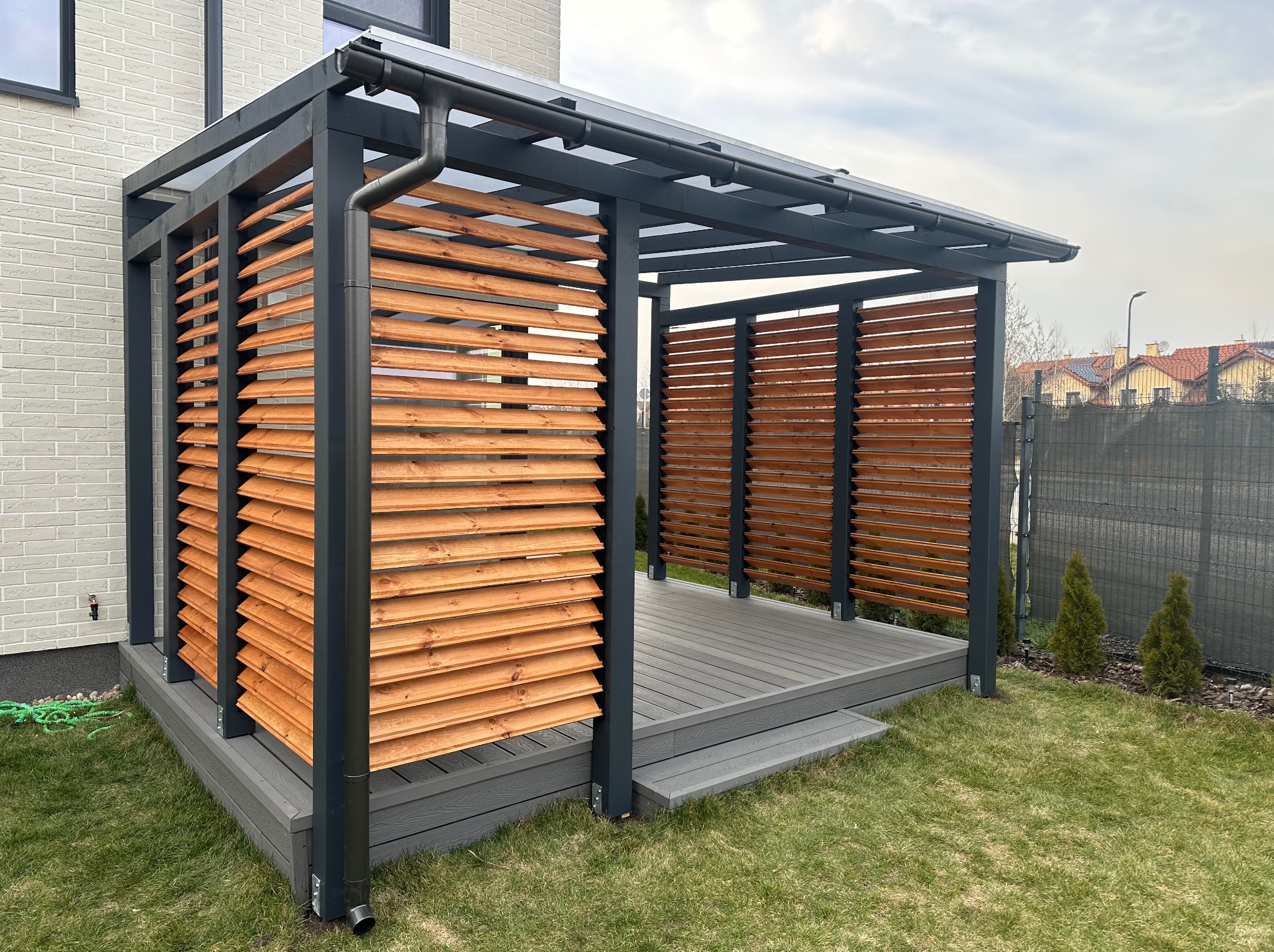 Nowoczesna pergola z drewnianymi lamelami i szarą konstrukcją, z widocznym systemem rynnowym, umiejscowiona na tarasie z desek kompozytowych, otoczona zielonym trawnikiem.
