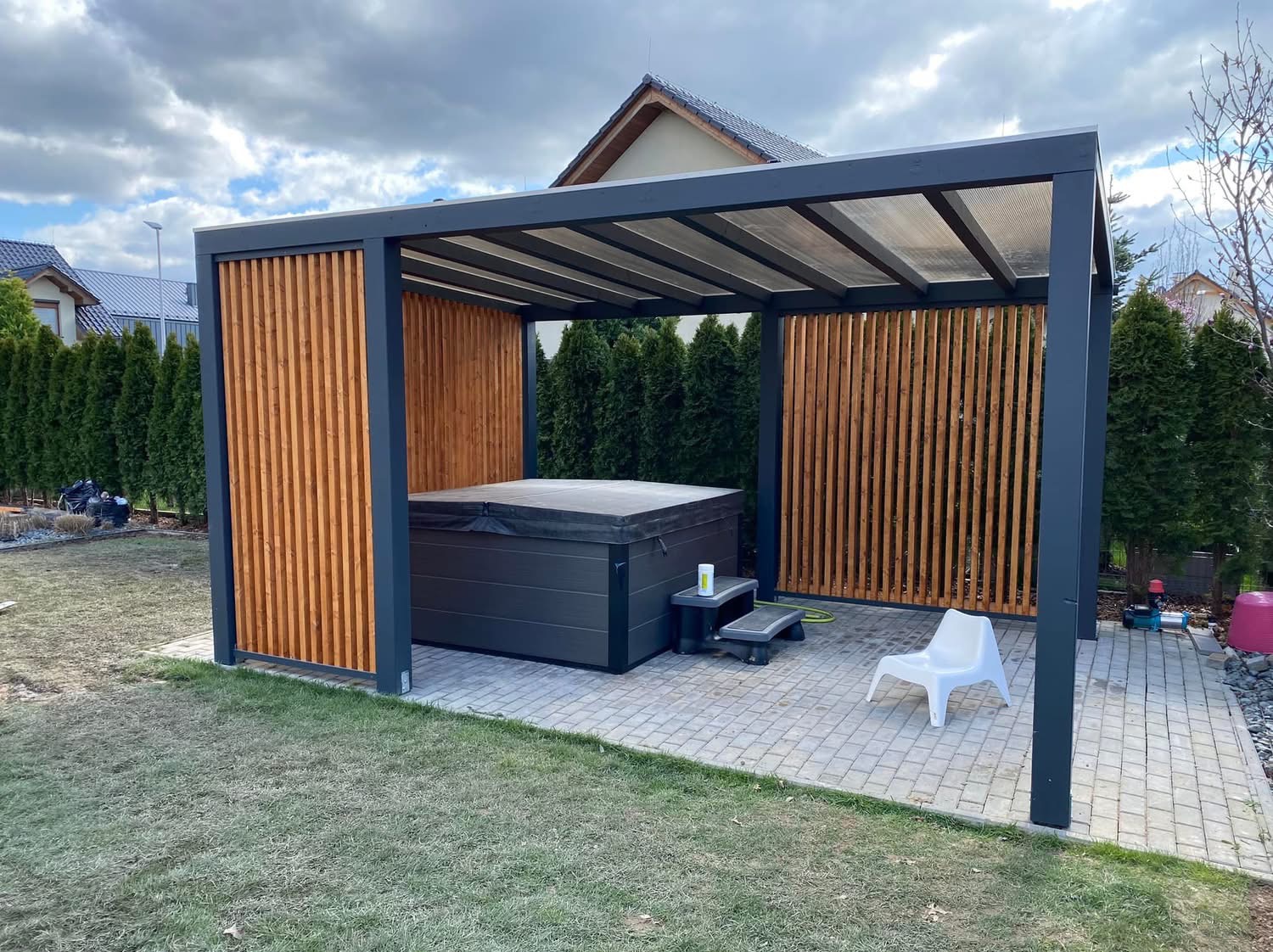 Nowoczesna pergola z drewnianymi panelami osłaniająca jacuzzi na kostce brukowej, otoczona trawnikiem i tujami.
