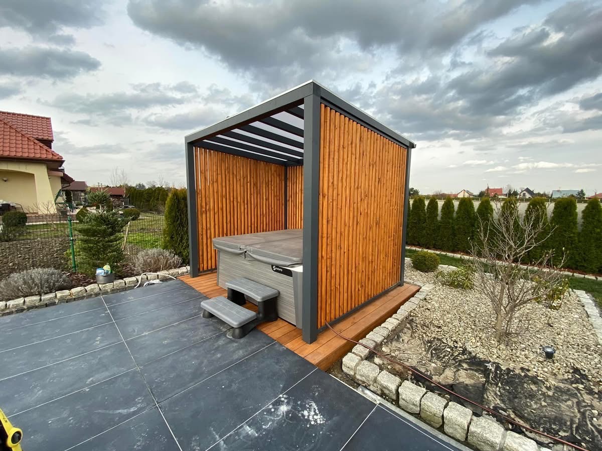 Nowoczesna pergola z drewnianymi panelami osłaniająca jacuzzi na tarasie z ciemnych płyt, widok na zadbany ogród i pochmurne niebo.