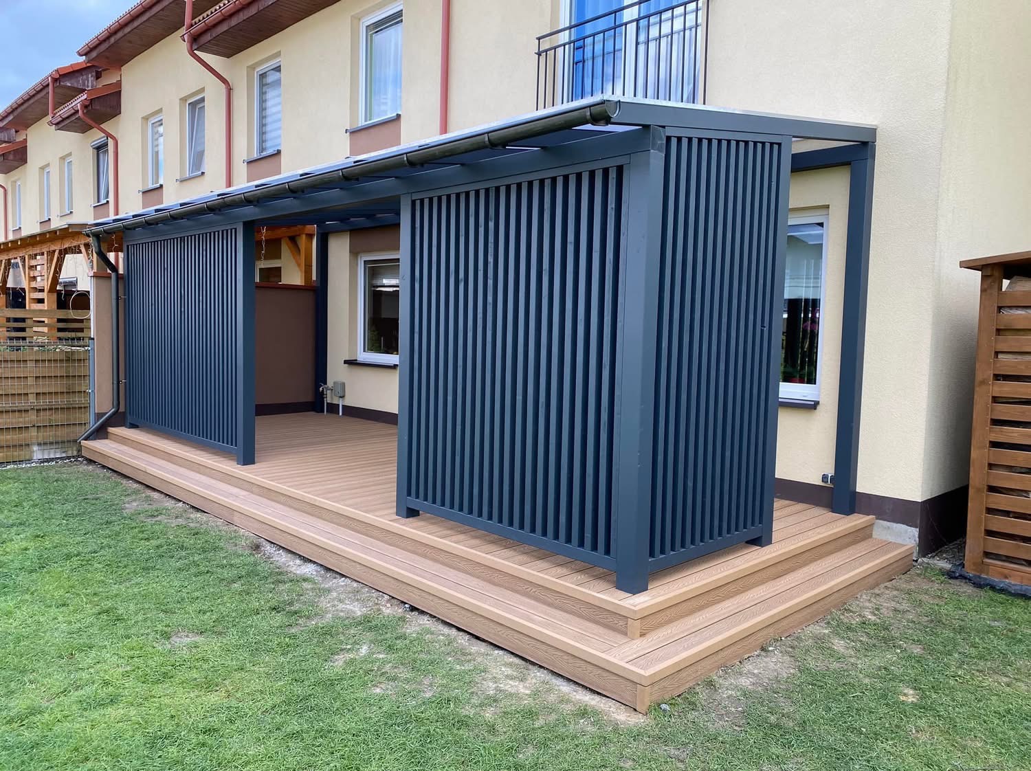 Zadaszony taras z kompozytu w kolorze jasnego drewna z ażurowymi, grafitowymi osłonami pionowymi, schody wejściowe z tego samego materiału, trawnik przed tarasem.