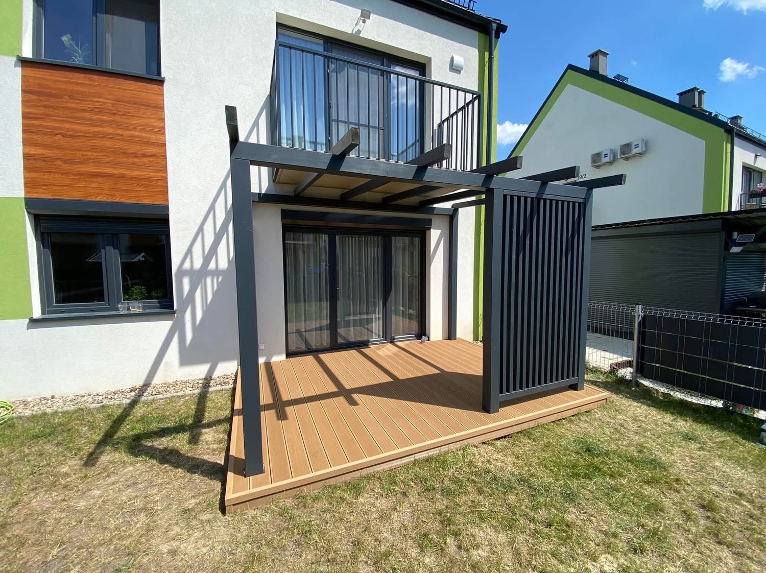 Nowoczesny taras kompozytowy z pergolą w kolorze antracytowym, przylegający do budynku z balkonem, widok z poziomu trawnika.