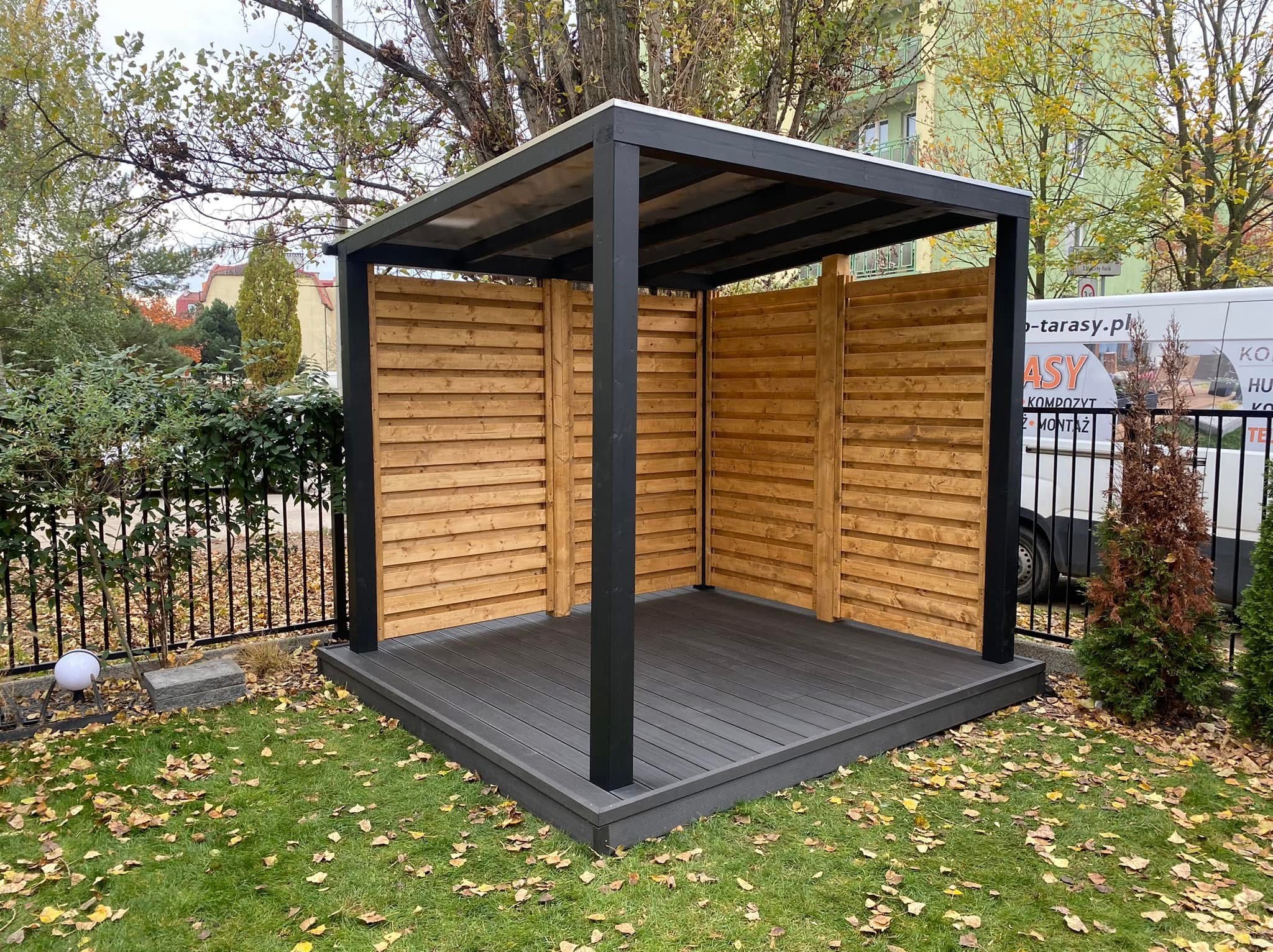 Nowoczesna pergola z drewnianymi panelami bocznymi i szarą kompozytową podłogą tarasową, widok z zewnątrz na tle zieleni i zabudowań miejskich.