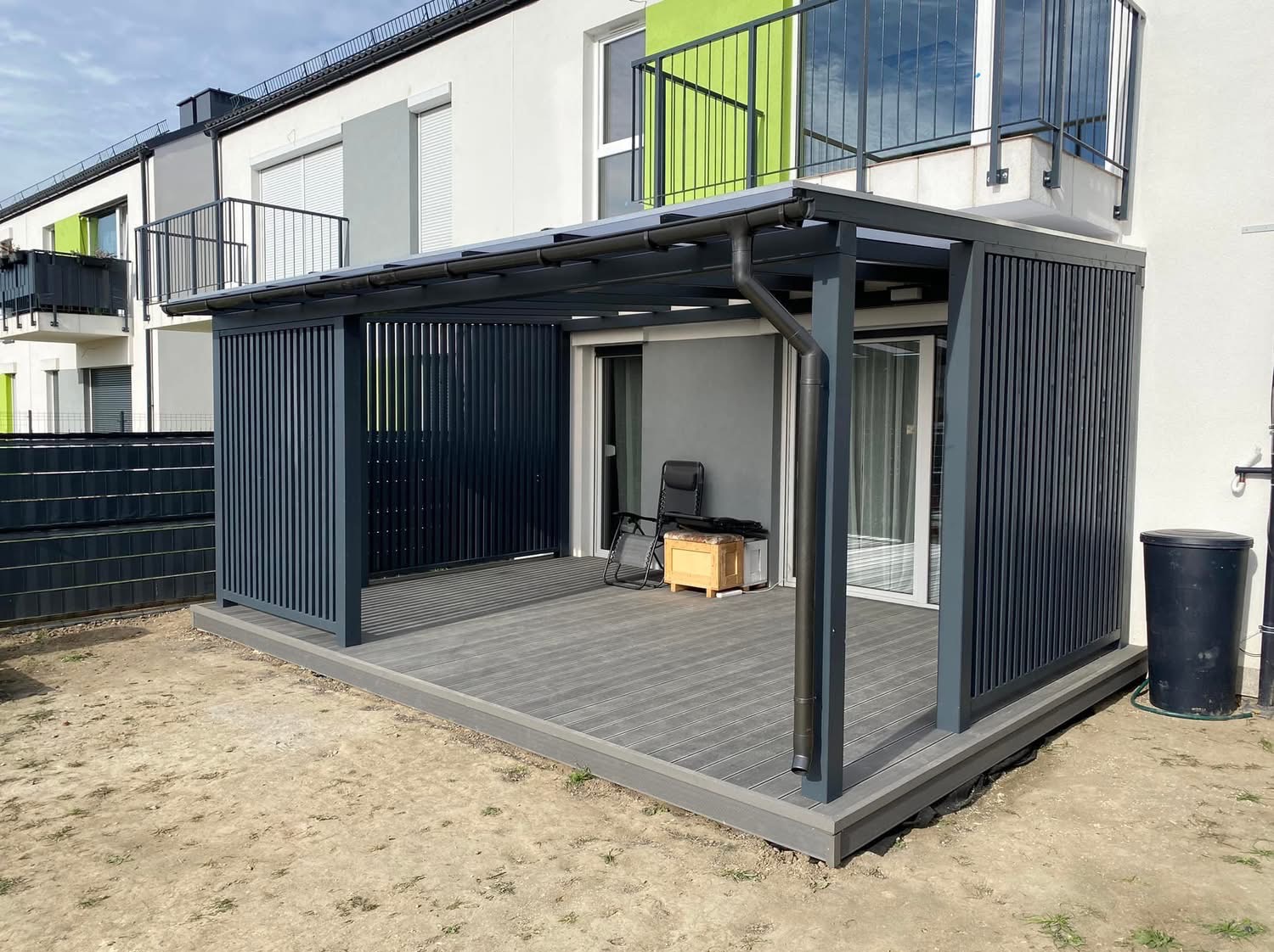 Nowoczesna, szara pergola z pionowymi panelami i zadaszeniem nad tarasem, z widocznym krzesłem i skrzynką, na tle budynku wielorodzinnego.