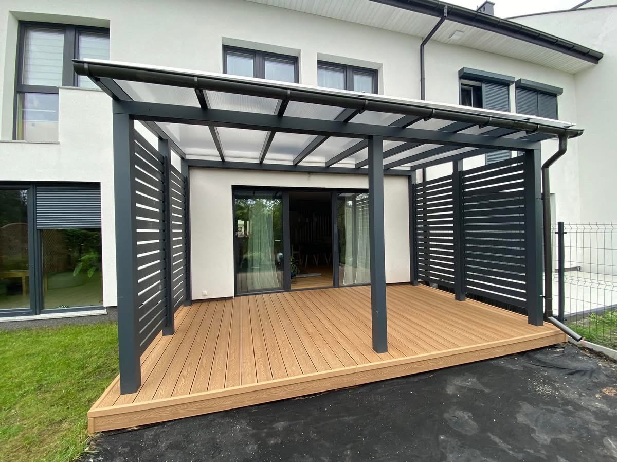 Nowoczesna pergola z szarymi, ażurowymi ściankami i przezroczystym zadaszeniem, połączona z tarasem z desek kompozytowych w kolorze jasnego drewna, przylegająca do białego budynku z dużymi oknami.