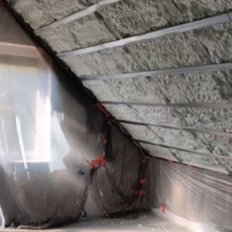 Poddasze z metalową konstrukcją i warstwą szarej pianki izolacyjnej, zabezpieczone folią ochronną przy ścianach.