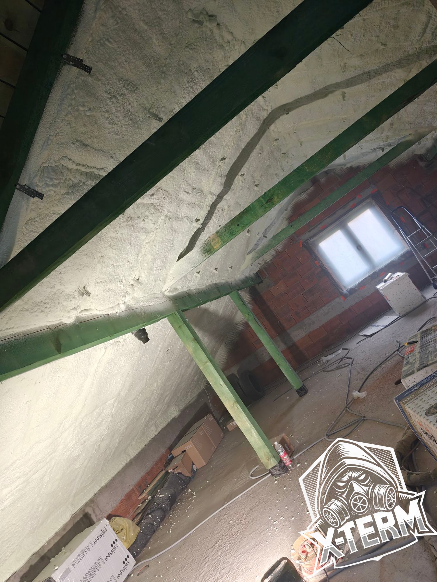 Poddasze z widoczną warstwą ocieplenia pianką poliuretanową, zielone drewniane belki konstrukcyjne, okno w murze z czerwonej cegły, częściowo widoczne logo firmy.