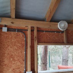 Ebaro Sp. z o.o. - Instalacja elektryczna w drewnianej altance: peszel przymocowany do ściany z płyty OSB, puszka elektryczna i włącznik światła, widok na drzewa przez okno.