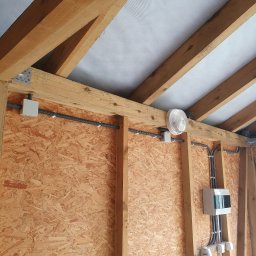 Ebaro Sp. z o.o. - Wnętrze konstrukcji drewnianej z widoczną rozdzielnicą elektryczną, kable w peszlach, gniazdo siłowe i lampa sufitowa, zamontowane na płycie OSB.