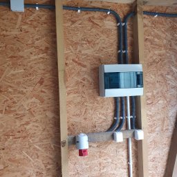 Ebaro Sp. z o.o. - Rozdzielnica elektryczna z przewodami w peszlach, zamocowana na ścianie z płyty OSB, widoczny także punkt oświetleniowy i gniazdo siłowe.