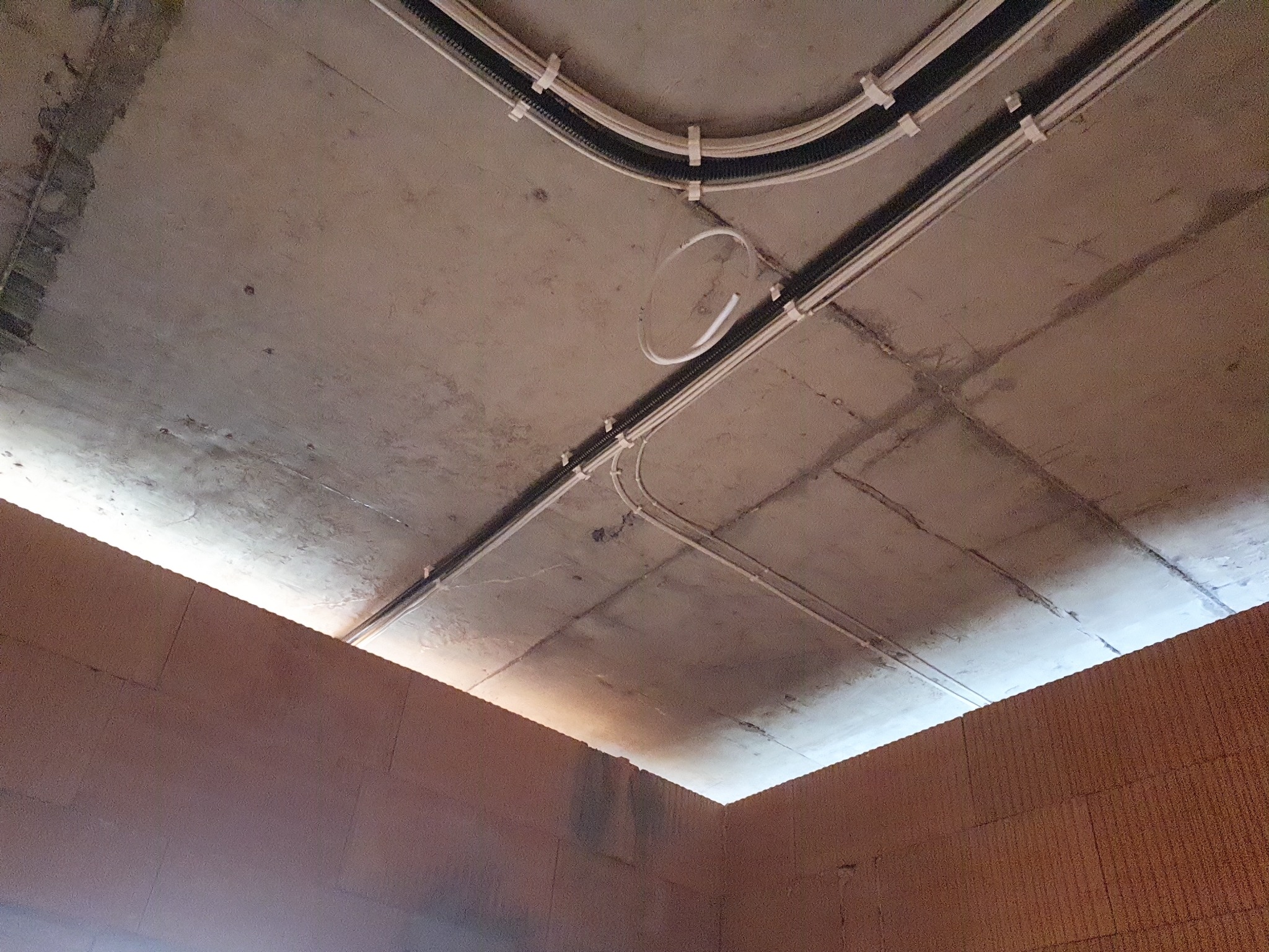 Betonowy sufit z zamontowanymi kablami w peszlach i rurkach, przymocowanymi do sufitu za pomocą uchwytów, widoczne także fragmenty muru z cegieł.