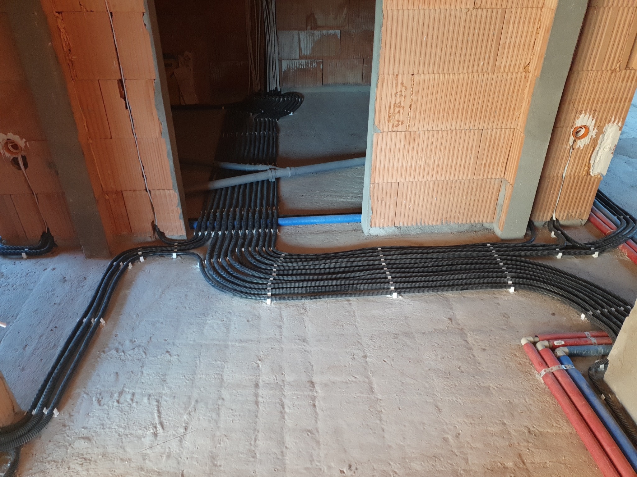 Rozprowadzenie peszli z kablami w surowym budynku z cegły, widoczne otwory instalacyjne w ścianach, kable ułożone równolegle i przymocowane do podłoża.