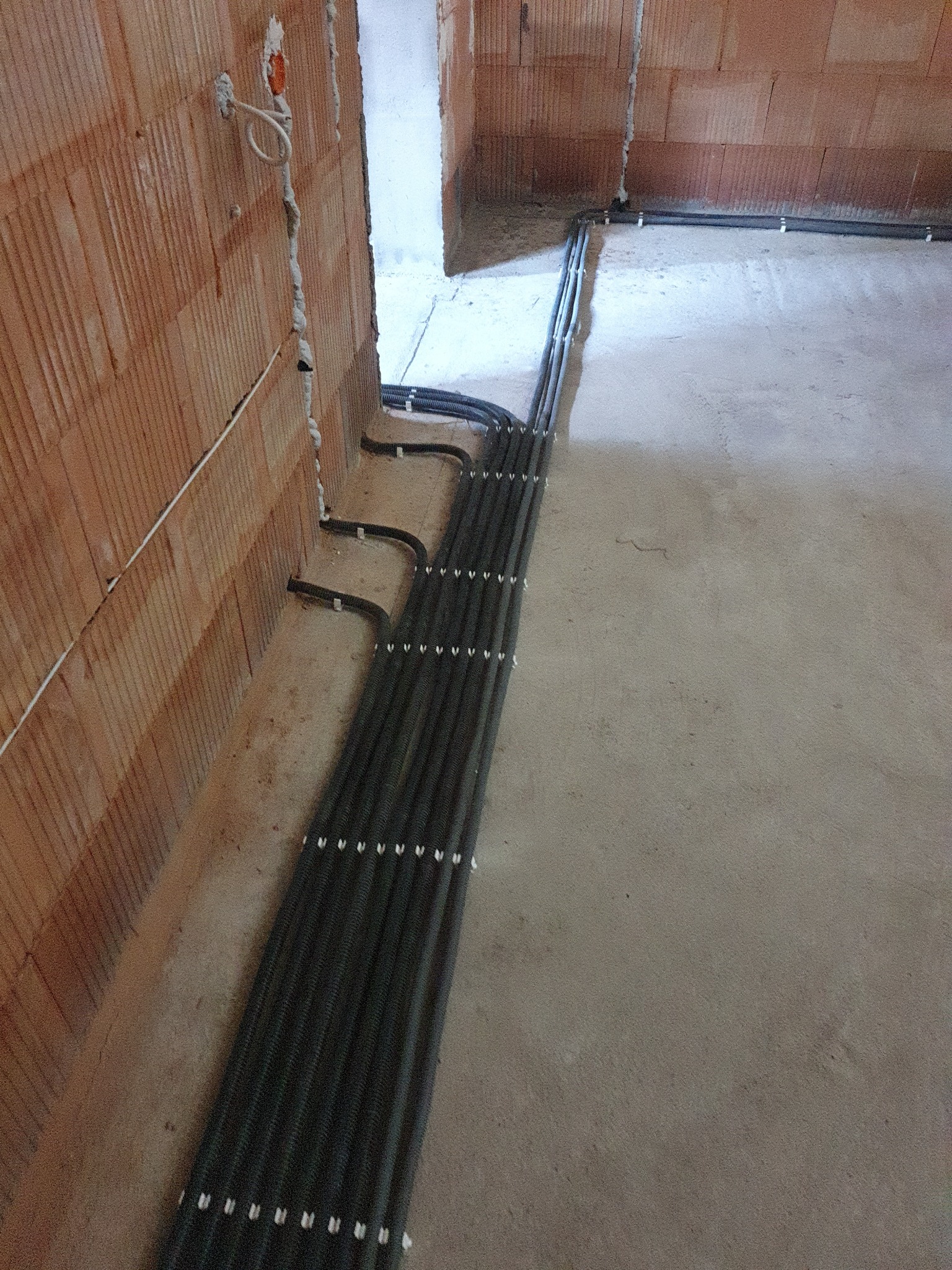 Wiązka przewodów elektrycznych w peszlach, prowadzona po podłodze w surowym stanie, mocowana do ściany z cegły dziurawki za pomocą uchwytów.