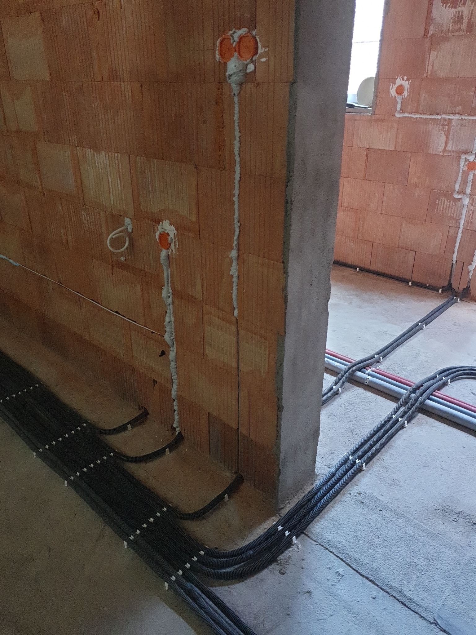 Instalacja elektryczna w trakcie montażu w surowym stanie: widoczne pomarańczowe puszki podtynkowe w ceglanej ścianie, kable w peszlach przypięte do podłoża, surowy betonowy filar w przejściu...