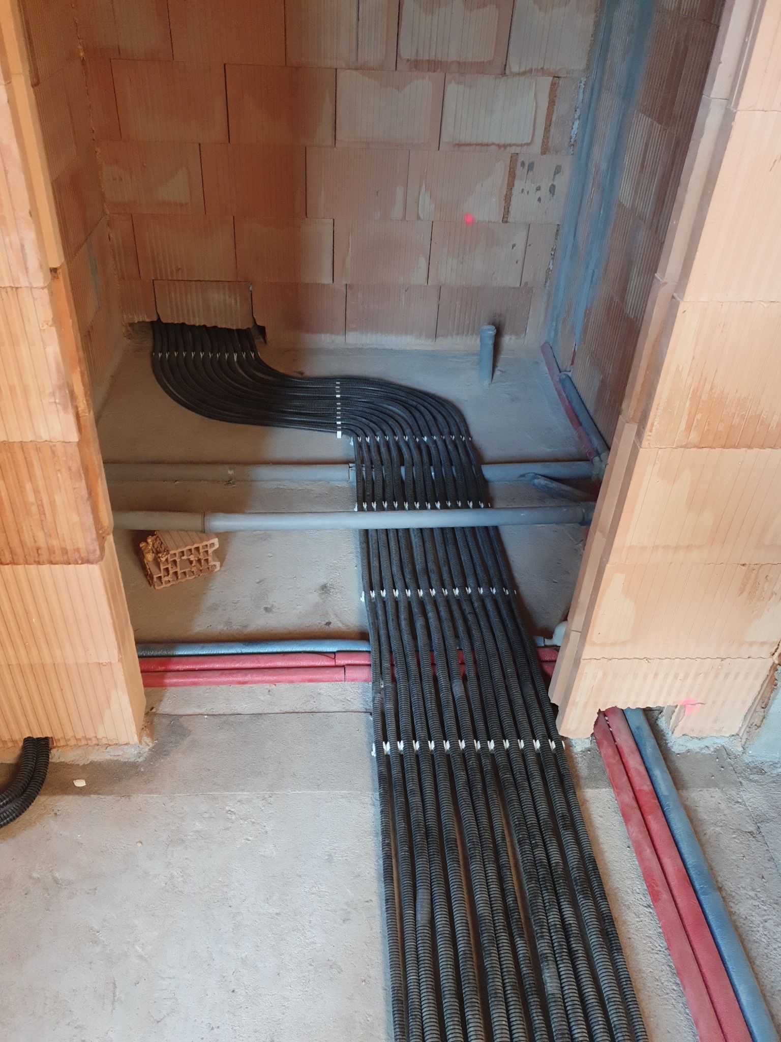 Wewnętrzna instalacja przewodów w karbowanych peszlach, ułożonych równolegle w surowym stanie budynku z cegły ceramicznej, widoczne rury wodne w kolorze czerwonym i niebieskim.