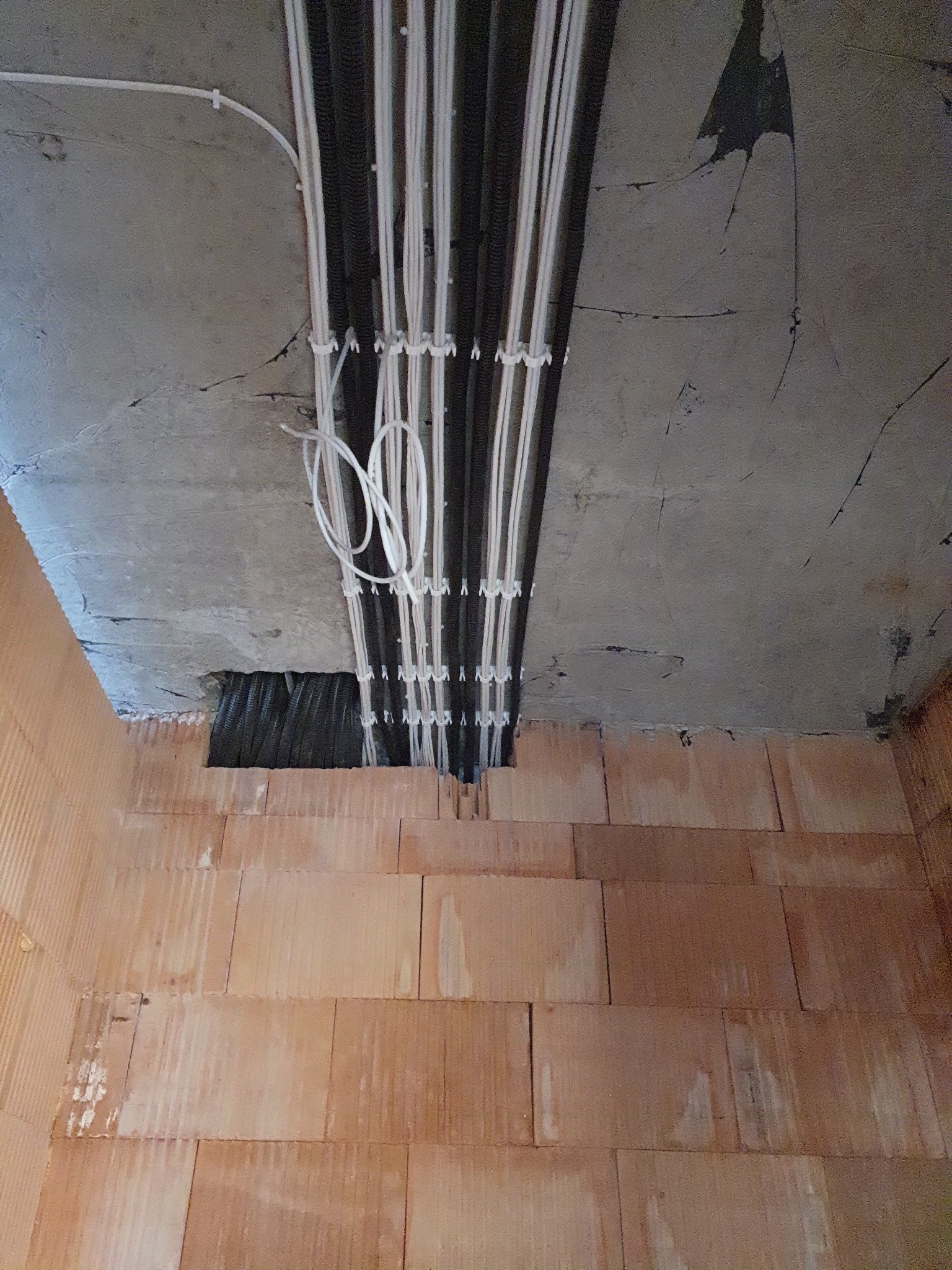 Wiązka przewodów elektrycznych w peszlach, częściowo białych i czarnych, przymocowanych do surowego betonowego stropu, w tle ściana z cegieł dziurawki.