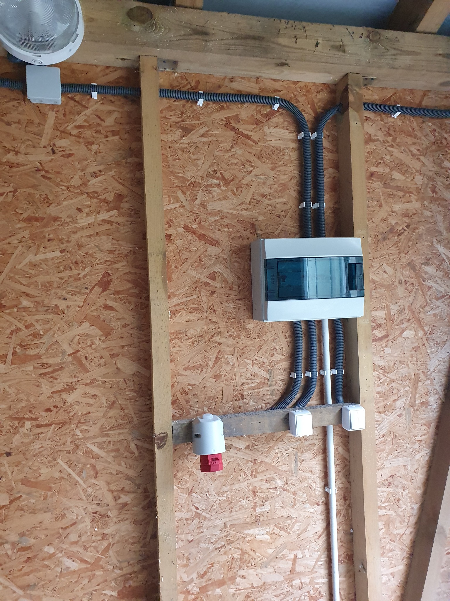 Rozdzielnica elektryczna z przewodami w peszlach, zamocowana na ścianie z płyty OSB, widoczny także punkt oświetleniowy i gniazdo siłowe.