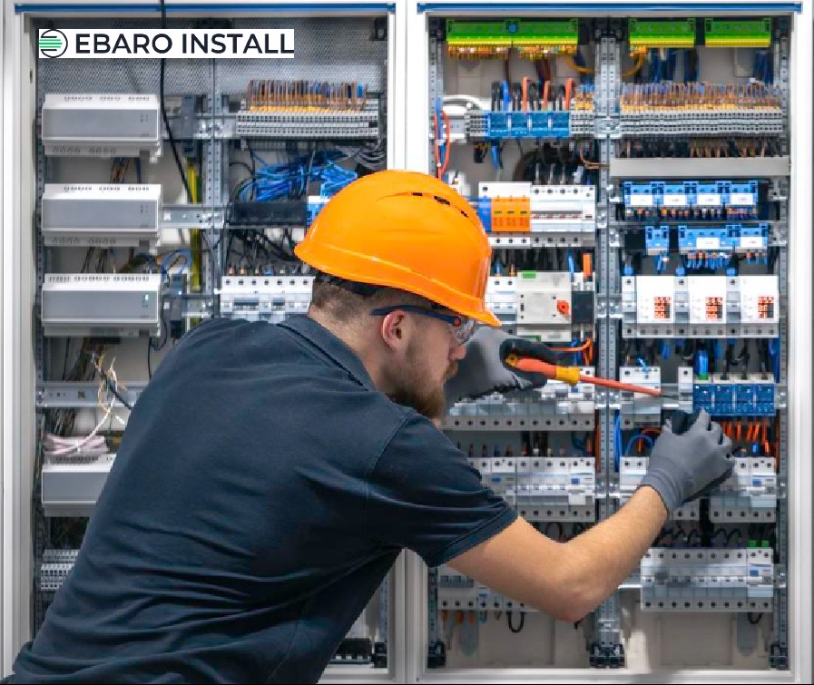 Elektryk w pomarańczowym kasku i okularach ochronnych, w szarych rękawicach, pracuje przy rozbudowanej szafie elektrycznej z bezpiecznikami i kablami, używając izolowanego śrubokrętu.