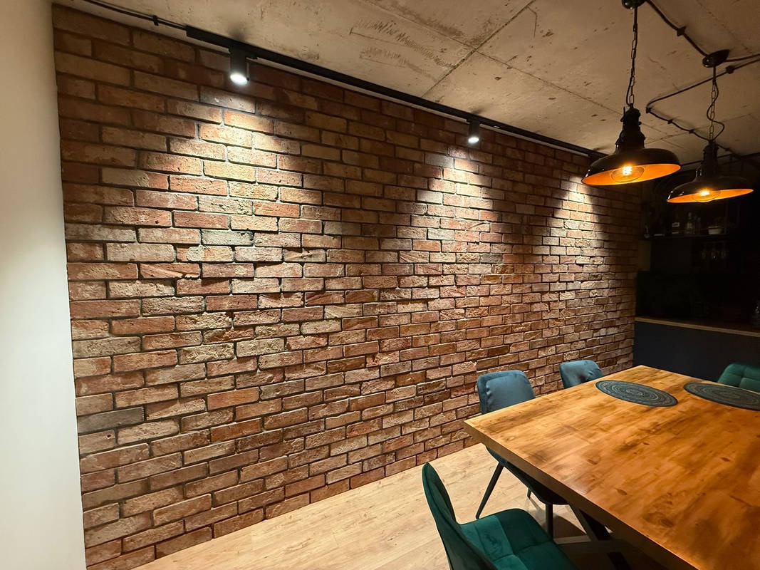 Ściana z cegły klinkierowej podświetlona reflektorami szynowymi, widoczny drewniany stół z krzesłami. Industrialny design wnętrza.