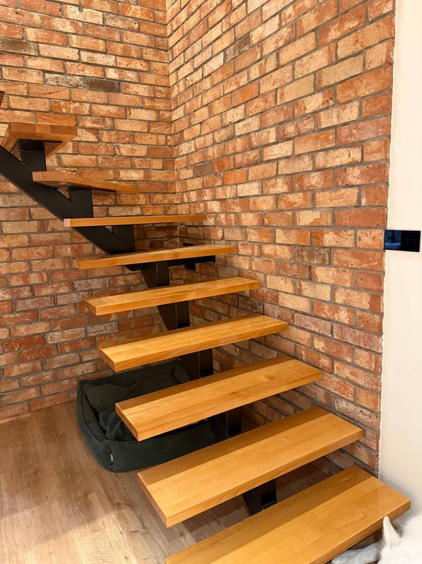 Drewniane schody wspornikowe z czarną metalową konstrukcją na tle ściany z cegły. Pod schodami leży zielona pufa. Widok z dołu, z bliska.