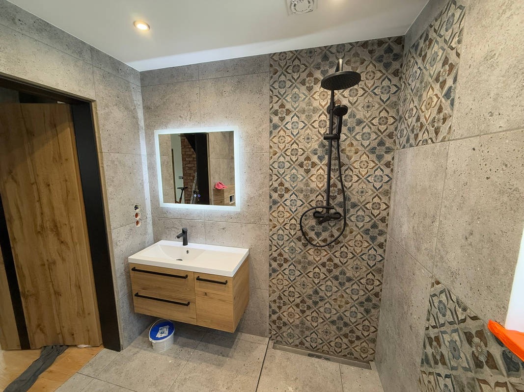 Wykończona łazienka z szarymi płytkami, umywalką z szafką z drewna, lustrem z podświetleniem LED i czarną armaturą prysznicową na ścianie z wzorzystymi płytkami.